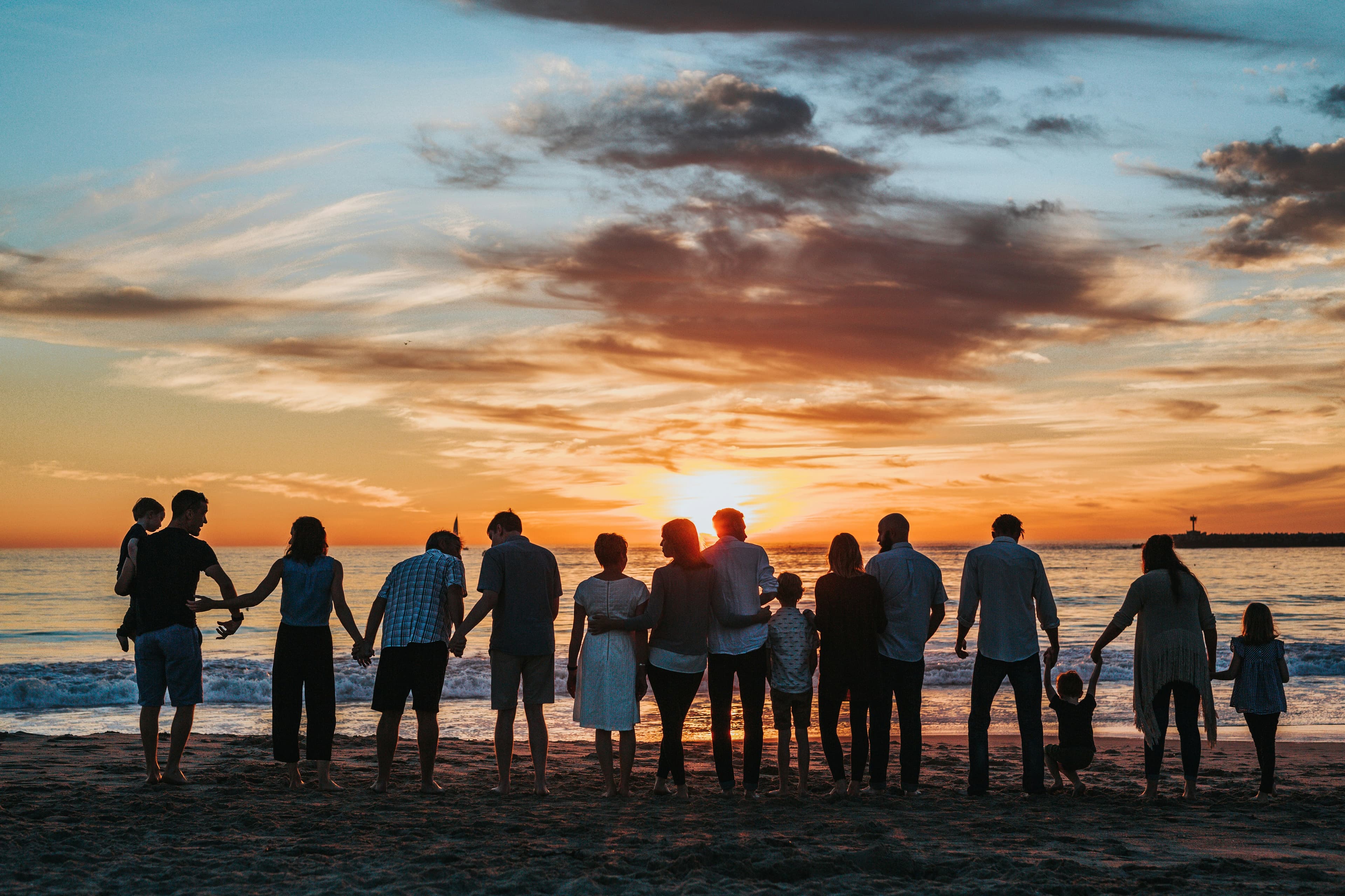 An extended family of adults and children silhouetted against a sunset beach, standing together holding hands